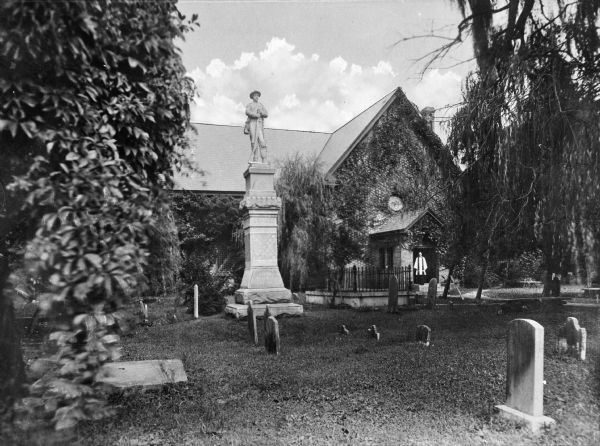 View of the church from the cemetery, which has a large monument. A clergyman stands in the doorway of the church. The building is brick, with vines trailing up the sides.