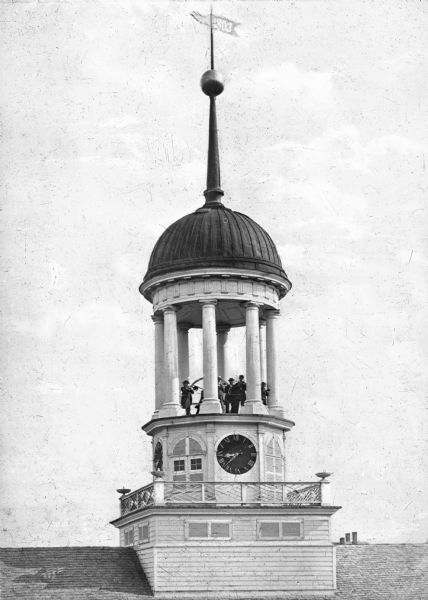View toward the round belfry on the roof supported by columns. A clock is below. A group of men pose around the bell, playing instruments.