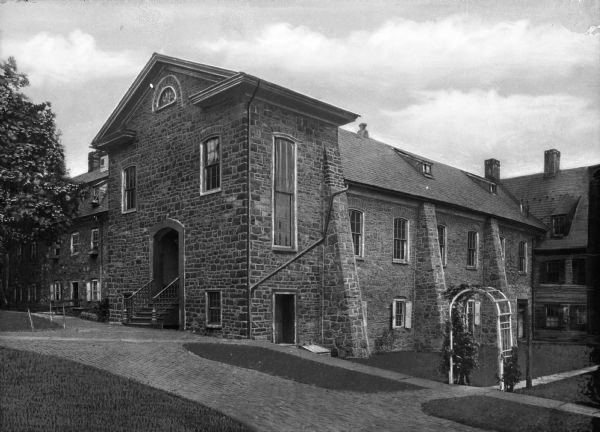 View across road toward the front and right side of the stone chapel. The facade has an arched entryway and three windows, and the side has rows of windows amongst the buttresses. There are buildings behind on the left and right, and an arched trellis entry to the walk bordered by a lawn is in the foreground at the sidewalk.