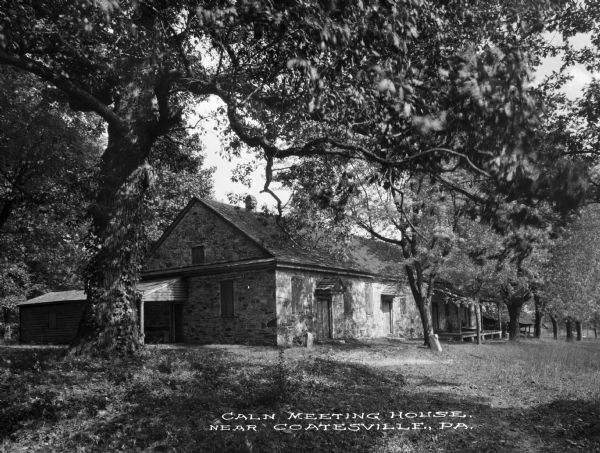 View across lawn with a large tree on the left toward the meeting house, which consists of three adjoining, one-story buildings. There are a number of doorways lining the front, with a porch on the end to the right. Connected to the structure is a wooden shed on the left. Caption reads: "Caln Meeting House, Near Coatesville, PA."