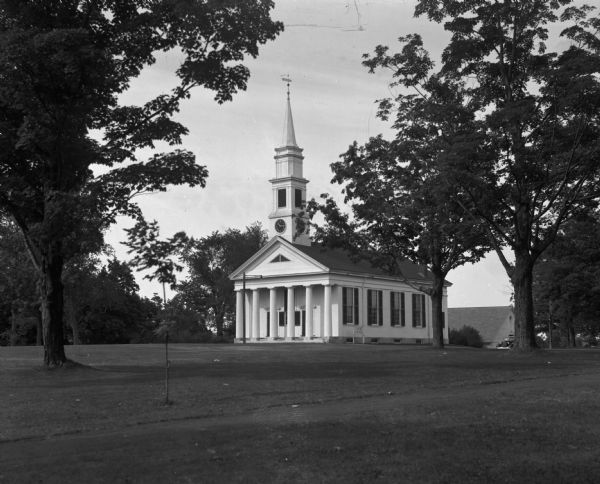 View across a broad lawn toward a church from a distance. The front has a porch with columns, along with two entrances. The right side has tall windows with wooden shutters. Below the steeple is a clock. A building is behind the church on the right.