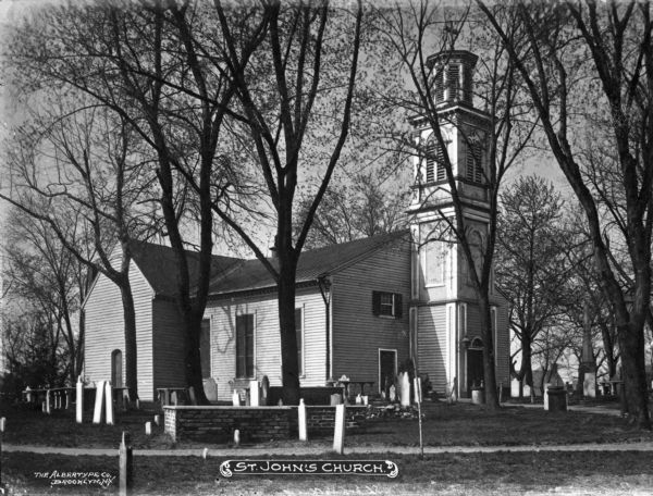 View from cemetery toward the church. The main entrance at the front is below the belfry. A path runs in front of the property. Caption reads: "St. John's Church."