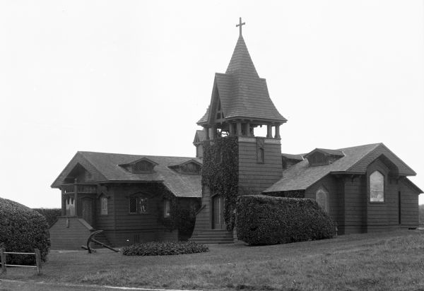 View across lawn toward the church. The wooden structure has two wings that form an L-shape, with a bell tower and main entrance in the elbow.  Vines cover the shingled walls. Some windows are multi-paned, and others are stained glass. An anchor is displayed in the yard.