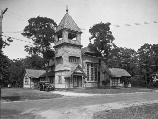 View across road toward a building with wooden siding and shingles, and a belltower above the main entrance. A man is posing in the automobile parked on the left of the building.