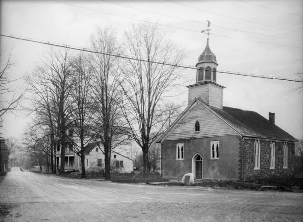 View from road toward the church. The windows have arched frames, and a set of double doors is on the front. Two houses are further down the street on the left.