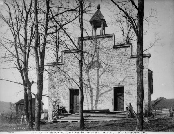 View toward the church's facade, which has a stepped roofline, and a bell tower on the roof. There are two entrances and a decorative railing framing the front steps. A hill is behind the church on the right, and a wooden building is on the left. Caption reads: "The Old Stone Church On The Hill, Everett, PA."