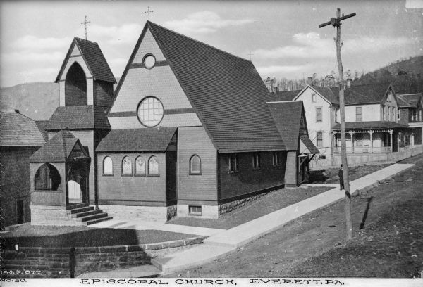 Elevated view of the church with a main entrance on the side. The building is covered in clapboard siding and shingles, with round and arched window,s and some with stained glass. In the background on the right is a house, with hills and trees in the background. A man stands on the sidewalk, but he is partially covered by the utility pole in the foreground. Caption reads: "Episcopal Church, Everett, PA."