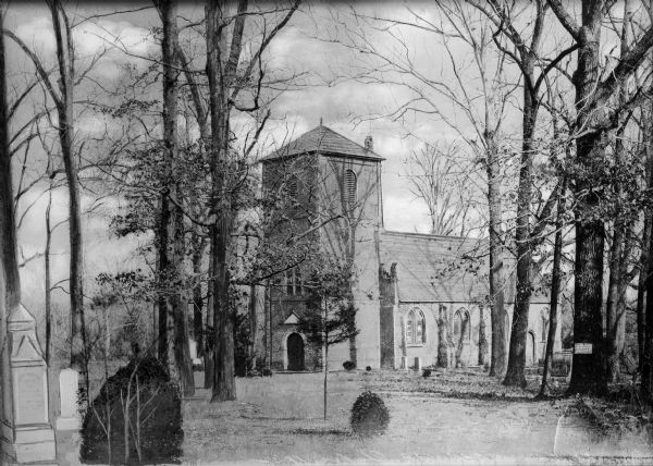 View from cemetery toward the brick church toward the front entrance, the belfry, and the right side. The side has arched, stained glass windows. Trees surround the property.