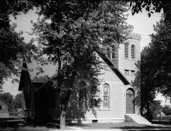 Exterior view of the stone church. The main entrance is below the belfry, and the arched window have stained glass. A man stands by a tree near the entrance. A sidewalk runs in front of the structure, with a path connecting the two.