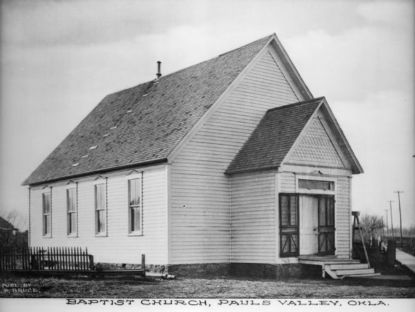 View toward the front and left side of the church which has wooden clapboard siding. The front entrance extends out from the building and has steps leading to a platform to the set of double doors. A picket fence is on the left. Caption reads: "Baptist Church, Pauls Valley, Okla."