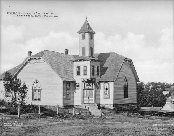 View toward the church. The decorative entrance is nestled in the corner between two of the wings of the structure. There are arched stained glass windows on the sides, and the windows in the belfry are plain and rectangular. Caption reads: "Christian Church, Chandler, Okla."