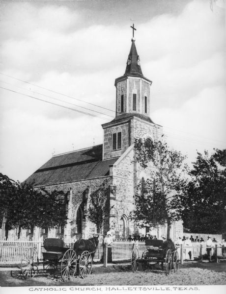 View across road toward the brick church with a fence along the sidewalk. There are men and women standing in the front yard. A picket fence runs around the property. Caption reads: "Catholic Church, Hallettsville, Texas."