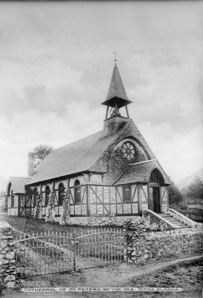 Elevated view across stone wall and gate toward the church with bell tower. The building is a stone structure with half timber walls, with the upper wall of the facade, and part of the roof covered in shingles. A rosette window is above the entrance. Hills and trees are in the background. Caption reads: "Cathedral of St. Peters-By-The-Sea, Sitka, Alaska." 
