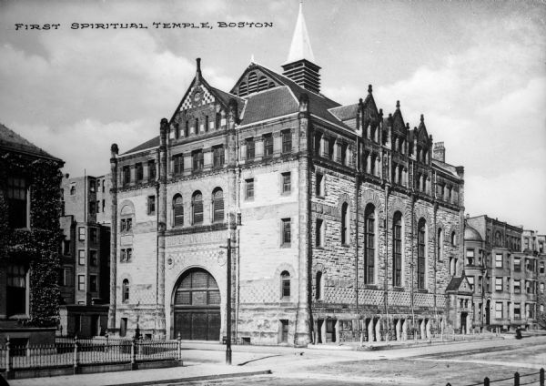 View across intersection toward the temple, which is a large stone building with a large arched entrance. Other structures and row houses are on the left and right. Caption reads: "First Spiritual Temple, Boston."