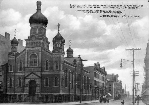 View down street toward the church, which is on the left corner with a fence surrounding the property. The structure is brick with stone detailing and has arched windows and doors. The windows are made of both regular and stained glass and the towers have onion domes. Men and carriages are in the street. Caption reads: "Little Russian Greek Catholic St. Peter and St. Paul Church, Corner Greene and Sussex Street, Jersey City, N. J."