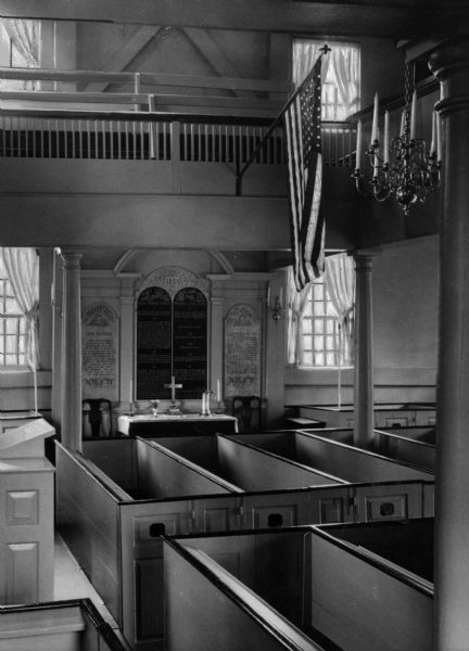 A view of the church's interior. A pulpit stands on the left, and on the back wall is an altar-like space, with a cross, candles, and a chalice. Hanging on the wall behind the altar are various prayers and Bible verses. In the foreground on the right are box pews, and a mezzanine seating area above with a flag on a pole on the front of the railing.