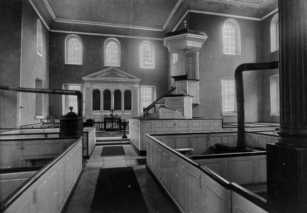 A view of the church's interior. The altar space on the back wall has an architectural feature on the wall that displays Bible verses. To the right is the elevated pulpit, with three different pulpits from which to speak. Various wood stoves and piping are among the boxed pews. Windows on two levels behind the altar and on the sides light the structure.