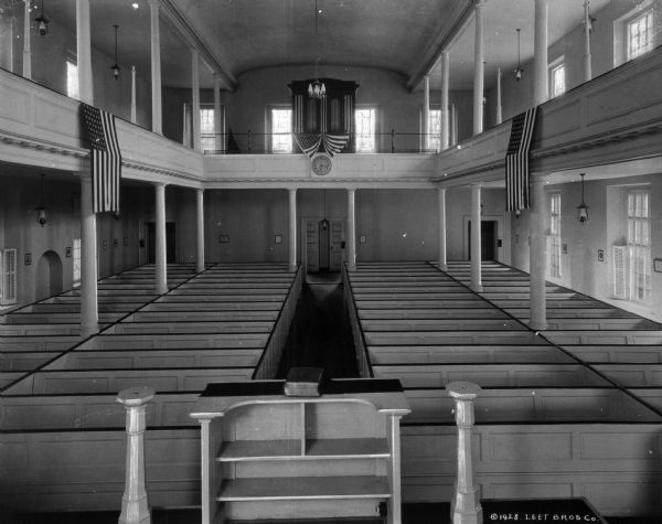 A view of the interior, taken from the altar, looking out to the seating area. U.S. flags hang from the mezzanine seating area, which also houses the organ. Plain windows light the interior.