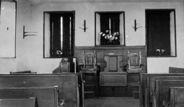 A view of the Stone Church interior looking toward the altar and first rows of pews. The altar consists of a raised platform with two chairs, each flanking the pulpit. The organ sits to the immediate left of the altar. Behind this area are three shuttered window openings, each with an arrangement of flowers on the sill.