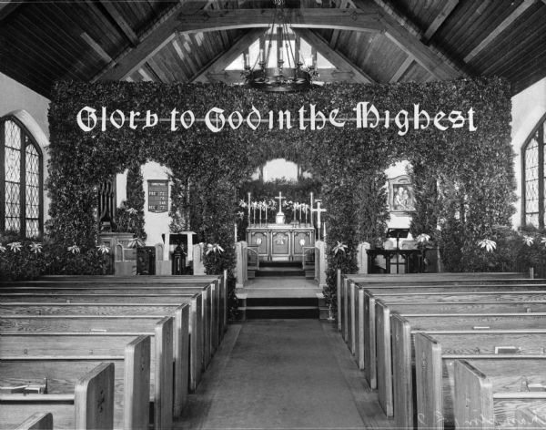 View down aisle toward the sanctuary area. An altar screen covered in vines separates the sanctuary area from the seating area and on in it is a banner that reads: "Glory to God in the Highest." To the left is the organ and pulpit, and to the right is a music stand. The altar area stands behind the screen and contains a crucifix, candles, and book.