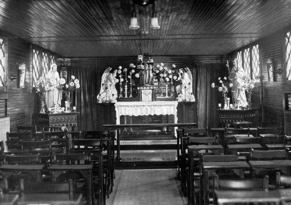 Chapel interior, showing the altar and seating areas. The altar has a lacy covering over the top. The back area contains the tabernacle, a crucifix, vases of flowers and candles. Statues on pedestals, both appearing to be angels, stand on either side. On the left is a side altar devoted to Mary with flowers and candles on it. The right side altar is devoted to Jesus and contains the same items as those found on Mary's altar.
