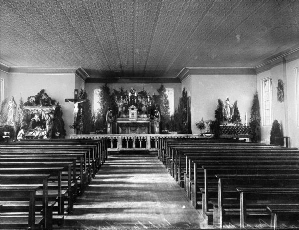 View down aisle toward the altar, with the pews decorated with evergreen trees and shrubbery. The ceiling has low relief moldings in a abstract pattern. The sanctuary has altar gates that separate it from the rest of the church. The altar sits in an alcove and is decorated with candles and an angel statue on either side. To the left of the alcove is a large crucifix, along with a side altar devoted to Jesus with a nativity scene. The right side altar is devoted to Mary.