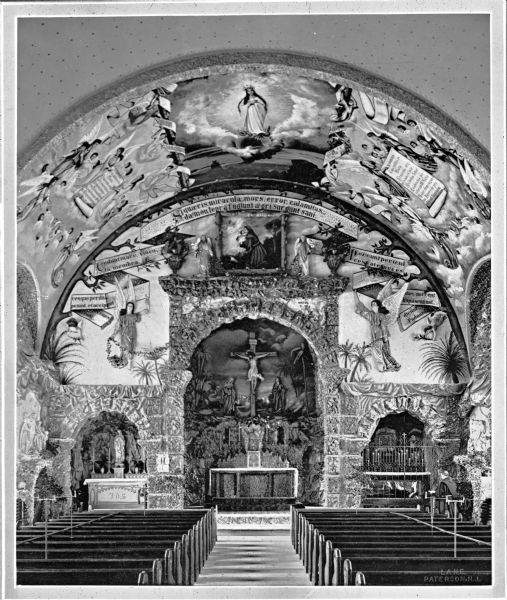 View down aisle toward the altar, and the murals surrounding the altar and on the arched ceiling at St. Anthony's church. The sanctuary consists of a main altar and two side altars, each with an arch highlighted in stonework. The altar has a covering over its surface, with a tabernacle in the middle; above this is a large crucifix. The left side altar is a grotto devoted to Mary. The right side altar contains two levels and has a tall metal grill in front of it. The upper level contains candles, and the lower level seems to contain an open coffin. A religious brother stands in front of this side altar with a book in hand.