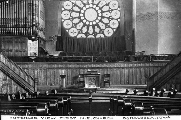 Interior view of First Methodist Church, including a rosette window, altar, and part of the seating area. The altar area contains a pulpit flanked by two chairs and tables with communion items stand nearby. On either side of the sanctuary are stairs leading to the choir area, containing chairs and a pipe organ. Caption reads: "Interior View First M.E. Church. Oskaloosa, Iowa."