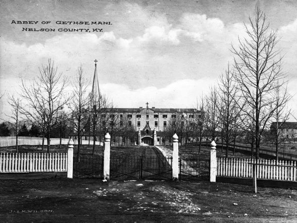 View of the abbey's facade, with the church's steeple visible behind and to the left of the monastery. A white picket fence with an iron gate entrance surrounds the property. The building's entrance is surrounded by a stone porch, and small windows line the upper floors. In the right background is a wood and stone structure. Caption reads: "Abbey of Gethsemani, Nelson County, KY."