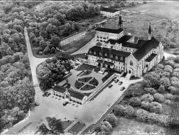 An aerial view of the monastery, showing a complex of buildings, roads, and the surrounding tree-covered property. A walled garden is between the entrance building and the living quarters. The church connects to the quarters.