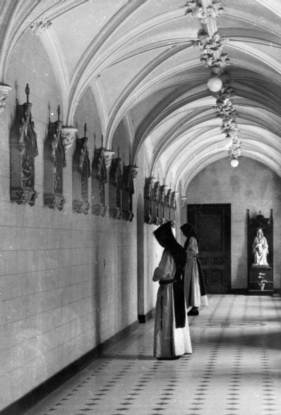 Two Trappist monks pray at the Stations of the Cross hanging on a hallway wall in abbey of Our Lady of the Valley. A statue of the Madonna and Child is sitting against the wall next to a door in the background.