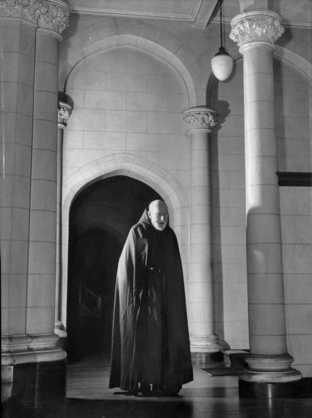 A Trappist monk in black robes stands in front of an arched doorway at the abbey of Our Lady of the Valley. The stone columns around him have floral capitals.