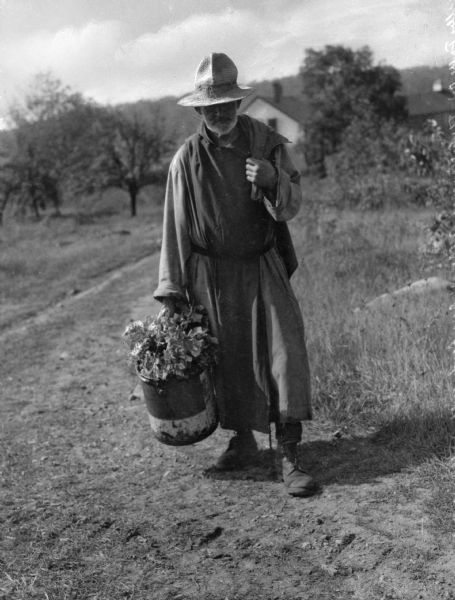Outdoor portrait of a Trappist monk holding a bucket of leaves or greens in one hand and carrying a sack over his opposite shoulder. Trees and a wooden structure are in the background.