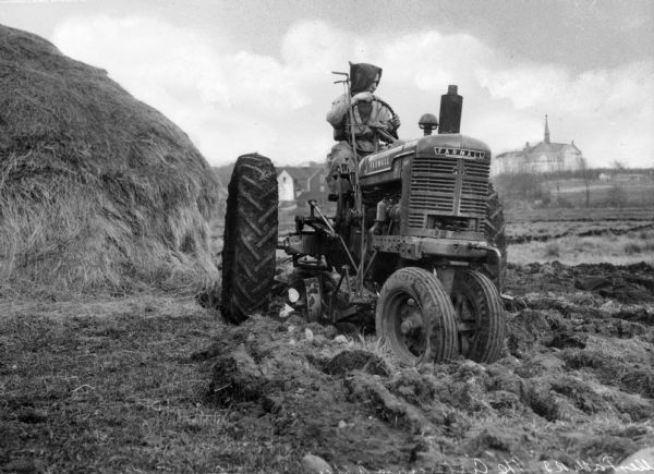View of a hooded Trappist monk on a tractor. A large pile of hay is on the left. The Our Lady of the Valley abbey and a wooden structure are in the background.