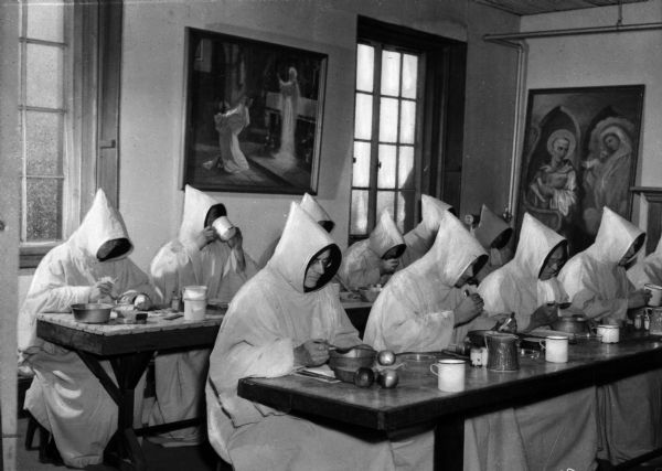 Trappist monks wearing white hooded cloaks sit at long tables while eating a meal. Religious imagery lines the walls.