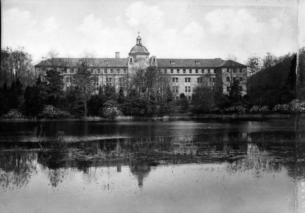 View of the Our Lady of the Cenacle convent from across a lake. The building stands among trees and has a main entrance area that juts out from the rest of the facade, which is topped by a dome and cross.