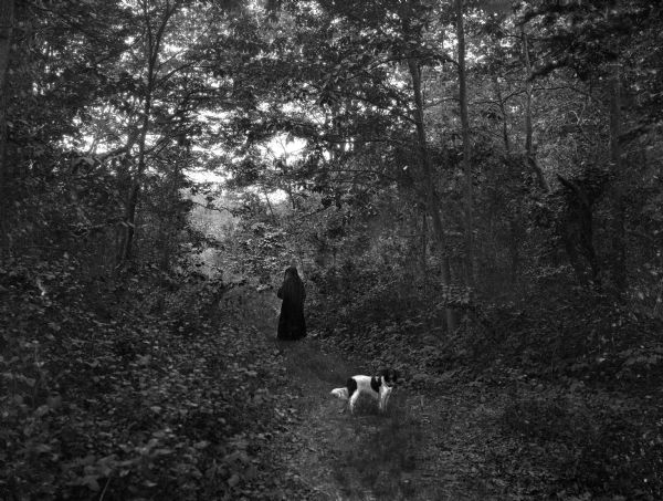 A nun, followed by a dog, walks down a path in the woods. The path is surrounded closely by trees.
