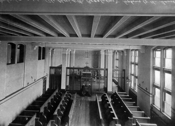 Elevated view of nuns kneeling in front of the sanctuary at Our Lady of the Cenacle, taken from the choir loft. Pews line the aisleway that leads to the sanctuary, which has an iron grill in front of it. Square columns are along both sides of the altar.