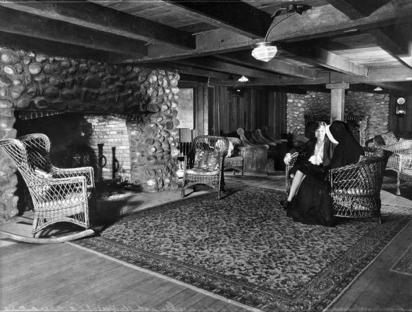 iew of a woman sitting with a nun in front of a fireplace in the Maude Adams guest house at Our Lady of the Cenacle convent. The room has low wooden ceilings, clusters of wicker furniture, and a large area rug in front of the fireplace.