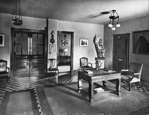 Interior view of a room in St. Joseph's guest house. A large wooden table and a chair are on the right, with a statue sitting on a pedestal in the back right corner.A set of double doors is on the left, and on the right is a doorway behind the table that leads to another room.