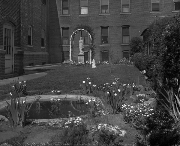 View of the Ave Maria Garden surrounded on two sides by a building. A large stone statue of Mary stands on a pedestal surrounded by an arched trellis; a smaller statue is in front of it on the right. Flowers and shrubs line the wall on the right, and a pond surrounded by flowers is in the foreground.