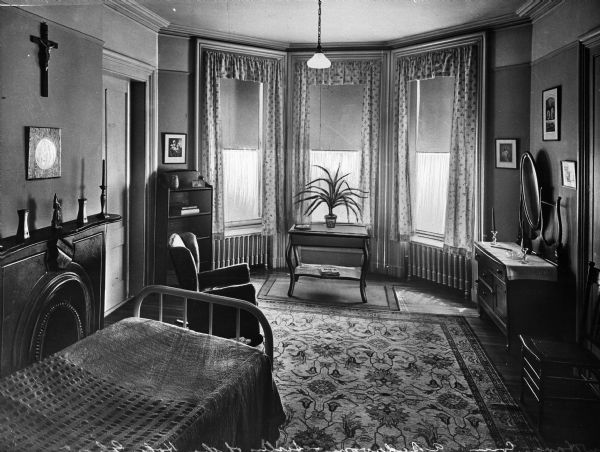Interior view of a bedroom at St. Joseph's guest house. The room is furnished with a bed, dresser, table, and chair. The fireplace on the left has an arched opening, and an area rug covers the floor.