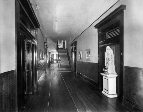 A view down the hallway at the School of the Brown County Ursulines. Mirrors hang on the walls and a statue stands to the right.  A staircase is in the background.