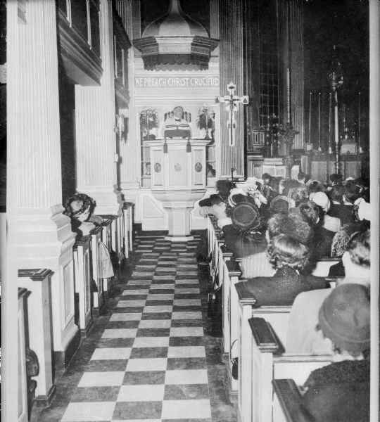 View down aisle toward a minister preaching from the elevated pulpit, while the congregation listens. To the right of the pulpit is the altar.