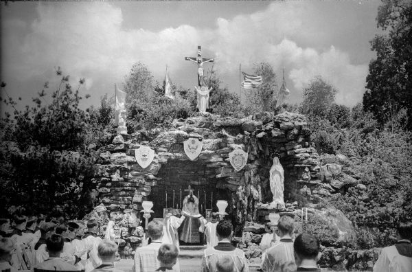 A view of a mass held at the grotto shrine on the Salesian grounds. The priest faces the altar, which is in a grotto, and a statue of Mary stands to the right. Statues and flags sit atop the grotto with a prominent crucifix in the middle. Shields with images on them hang from the entrance. A group of young men is in the foreground.