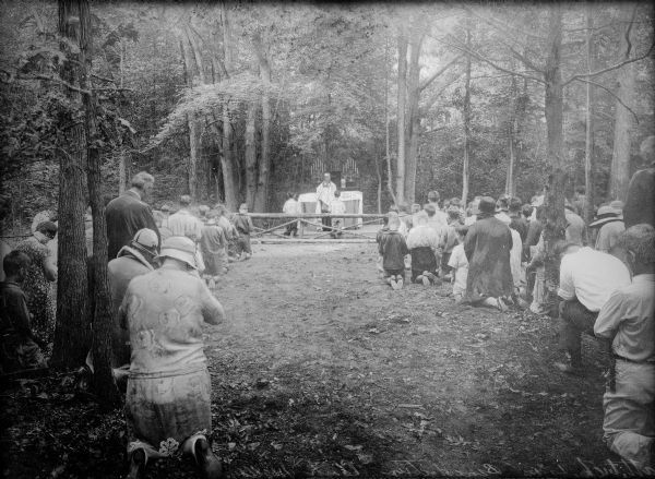A view of a benediction service in the woods at Camp Malloy. Men, women, and children stand or kneel on the ground beneath trees, while a priest stands at an altar in the background.