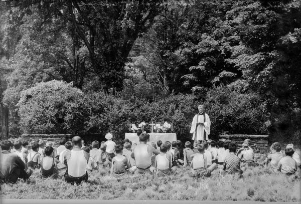 A clergyman performs an outdoor church service to boys of various ages.  A small stone wall flanks each side of the altar area. The altar contains flowers, a candle, and a crucifix.