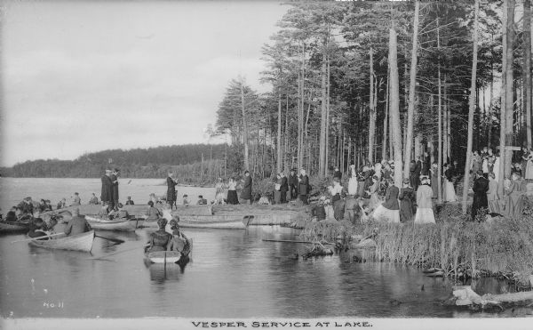 View across water along shoreline toward a Vesper service held by a lake. Clergymen stand on the dock while members of the congregation stand on the shore or sit in rowboats. Caption reads: "Vesper Service at Lake."