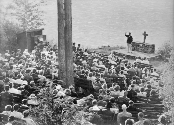 Elevated view of Vesper Services. In the background near the shoreline the minister stands near an altar on a stone platform to perform a vesper service. A crucifix sits atop the altar and an organ is to the left. In the foreground people sit for the service on wooden benches grouped in a half-circle around the platform.
