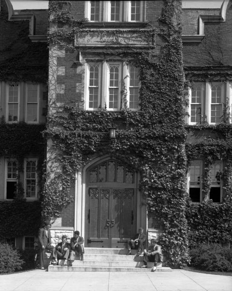 View of male students sitting on the entrance steps of a Princeton University campus building. The door and windows are arched and reflect a Collegiate Gothic style.  Ivy climbs the sides of the stone-trimmed brick building and partially obscures a sign above the door reading, "Princeton Class...J.D. Dormitory...1903(?)".